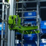 <h1>The Future of Logistics: Implementing a Provably-Safe Autonomous Compiler</h1> High-tech automated warehouse system featuring a green robotic arm handling blue storage crates.