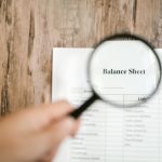 The Hidden Balance Sheet: Mental Health as Economic Capital Close-up of a balance sheet document on wooden surface with a magnifying glass held by a hand.