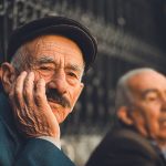 Elderly man in thoughtful pose wearing a cap, sitting outdoors in a sunny setting.