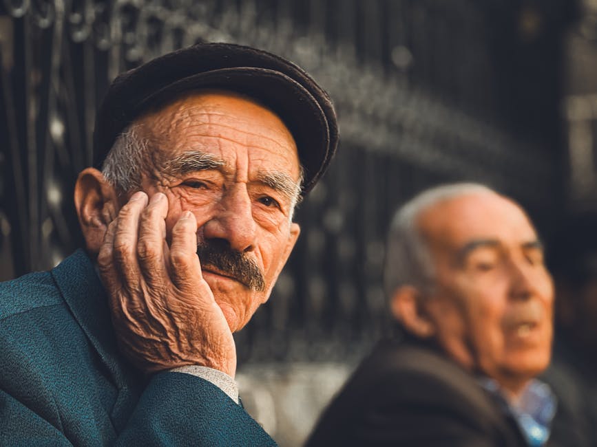 Elderly man in thoughtful pose wearing a cap, sitting outdoors in a sunny setting.