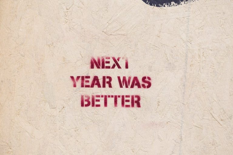 Rustic wall art with an inspirational message 'NEXT YEAR WAS BETTER' stenciled on a textured surface.