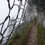 A solitary hiker walks along a misty mountain trail secured with protective netting.