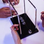 The Physics of Cities: Applying Systems Science to Urban Design Close-up of hands interacting with a Newton's cradle on a white table in a studio setting.