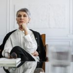 The Psychology of Style: Executive Presence and Strategic Identity A thoughtful professional woman sits at a glass table in a well-designed office, conveying contemplation and expertise.