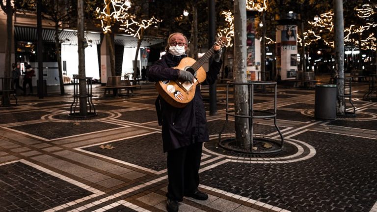 Elderly man playing guitar outdoors at night, surrounded by festive lights. Celebrating music and resilience.