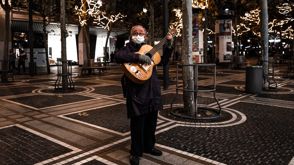 Elderly man playing guitar outdoors at night, surrounded by festive lights. Celebrating music and resilience.