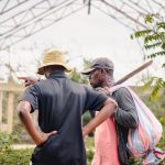 A group of three adults talking outdoors with greenery, one carrying a machete.