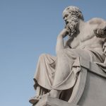 The Stoic Operator: Why Philosophy Is the Next Frontier of Performance Stone statue of philosopher Socrates sitting in thoughtful pose in Athens, Greece under a clear blue sky.