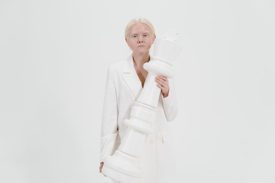 Woman in white outfit holding a large white chess king against a plain background.