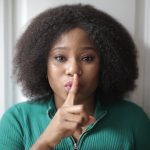 The Strategic Silence: Privacy as a Tool for Intellectual Sovereignty Portrait of a woman with afro hair signaling silence by holding a finger to her lips.