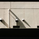 The Surveillance Paradox: Balancing Oversight and Ethical Leadership A minimalist wall featuring multiple security cameras casting long shadows in the sunlight.