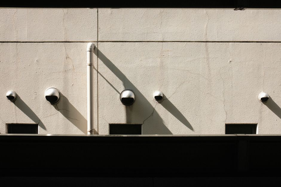 A minimalist wall featuring multiple security cameras casting long shadows in the sunlight.