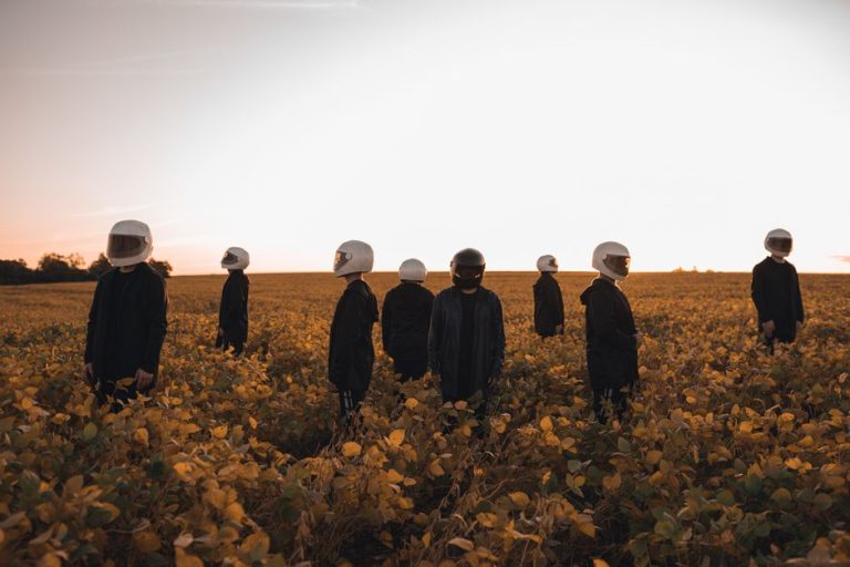 A group of people in astronaut helmets standing in a field of yellow flowers at sunset.