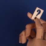 Uncertainty-Quantified EdTech: Post-von Neumann Systems Wooden mannequin hand holding a block with a question mark against a blue background.