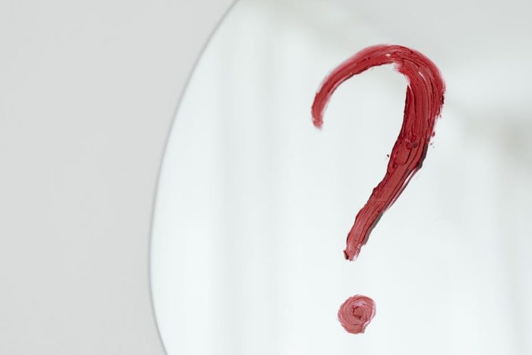 Close-up of a red question mark painted on a mirror with a white background.