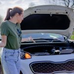 A woman looks puzzled by her broken car with an open hood on a rural road.