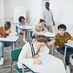 Group of students taking a test in a modern college classroom under teacher supervision.