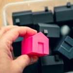 Hand selecting a pink house model from a box of miniature homes, symbolizing real estate choices.