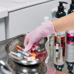 Close-up of a cosmetologist's gloved hand preparing syringes on a tray in a clinic setting.