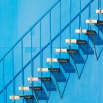 A minimalist view of a steel staircase casting shadows on a blue wall in sunlight.
