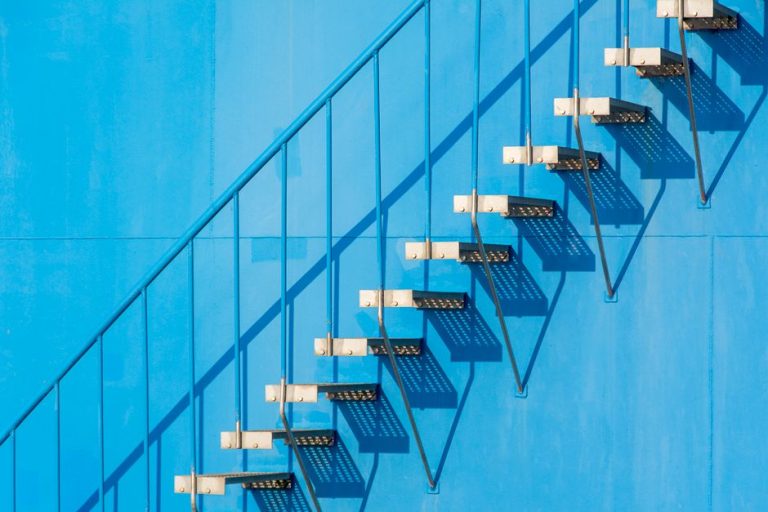A minimalist view of a steel staircase casting shadows on a blue wall in sunlight.