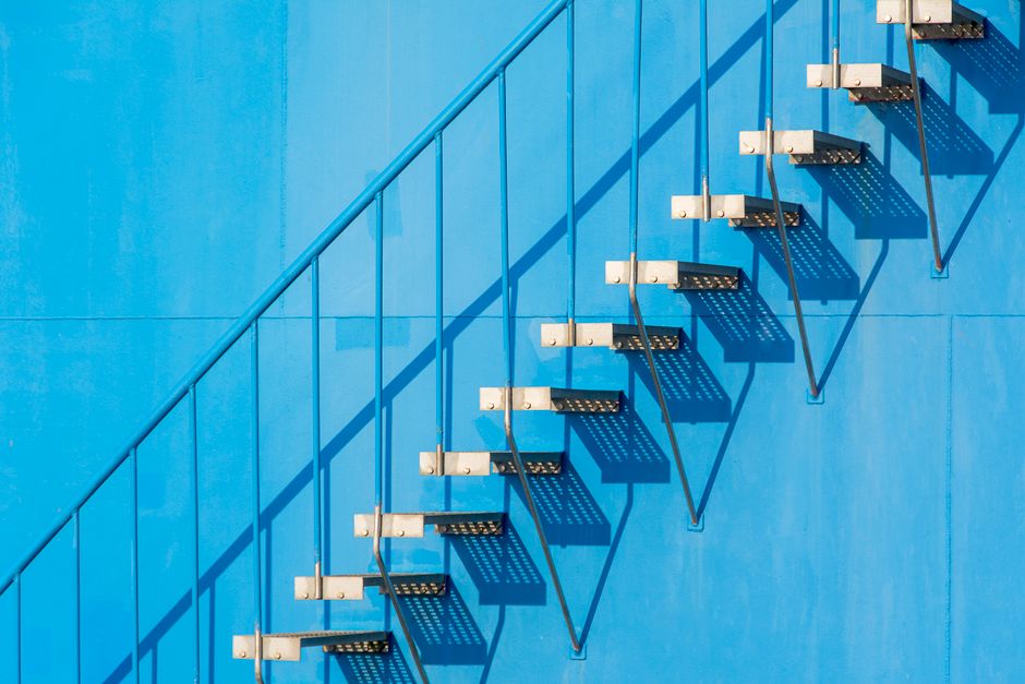 A minimalist view of a steel staircase casting shadows on a blue wall in sunlight.