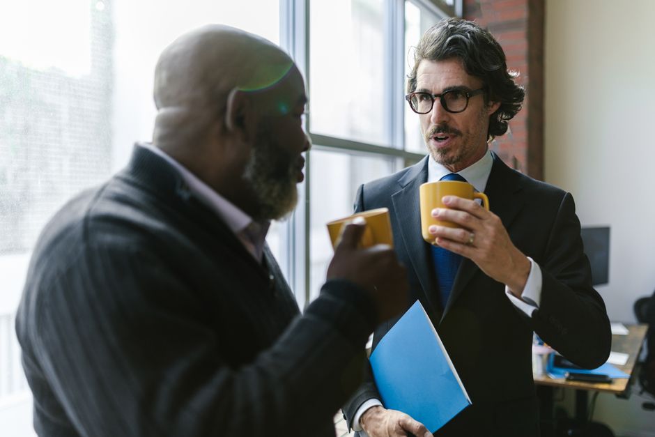 Two diverse professionals in corporate attire having a casual conversation over coffee indoors.
