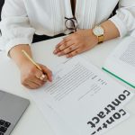 Close-up of a businesswoman signing a contract at her desk, with a laptop and book.