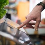 Automated Enforcement: Scaling Operational Excellence in 2026 A hand interacting with a digital weighing scale in a produce section, emphasizing technology in shopping.