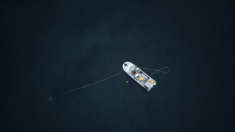 Aerial photograph of a single boat anchored in a vast, dark ocean, captured from above.