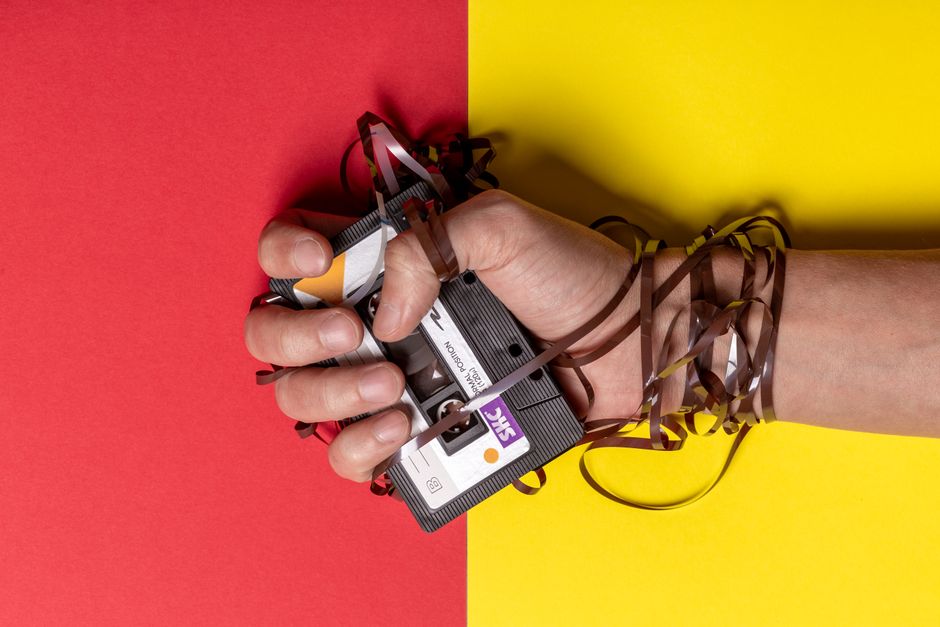 Hand holding tangled cassette tape on vibrant red and yellow background, representing nostalgia.