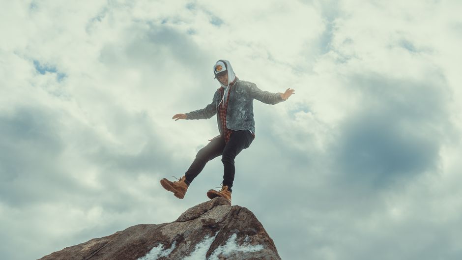 A man in casual attire balancing on a rock with a cloudy sky as a backdrop, exuding adventure.