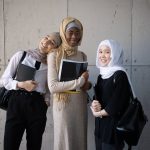 Delighted multiethnic female students in hijabs looking at camera while standing with backpacks and copybooks in hands near wall in corridor of university