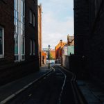 A narrow alleyway flanked by rustic brick buildings, leading to a sunlit street beyond.