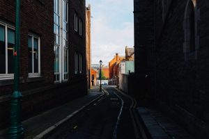 A narrow alleyway flanked by rustic brick buildings, leading to a sunlit street beyond.