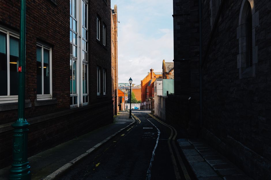 A narrow alleyway flanked by rustic brick buildings, leading to a sunlit street beyond.