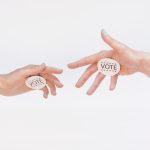 Two hands exchanging vote stickers symbolizing political participation and civic duty.