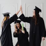 Three young women celebrating graduation with a high five, holding diplomas indoors.