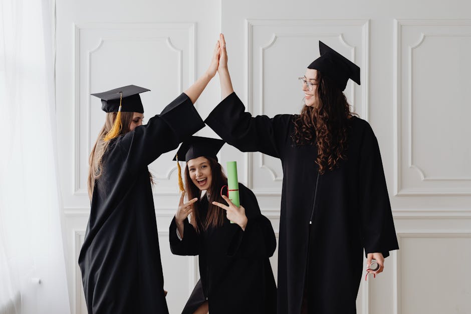 Three young women celebrating graduation with a high five, holding diplomas indoors.