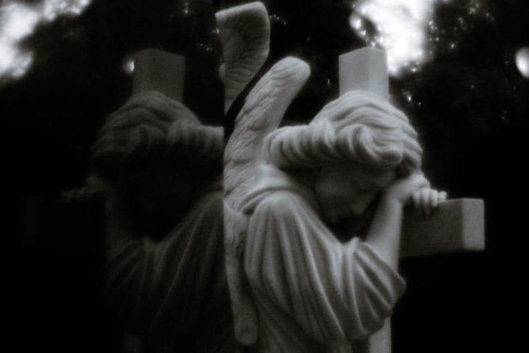 A reflective black and white photo of a solemn angel statue embracing a cross, evoking spirituality.