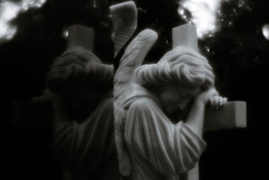A reflective black and white photo of a solemn angel statue embracing a cross, evoking spirituality.