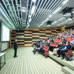 A speaker delivers a presentation to an attentive audience in a modern auditorium setting.