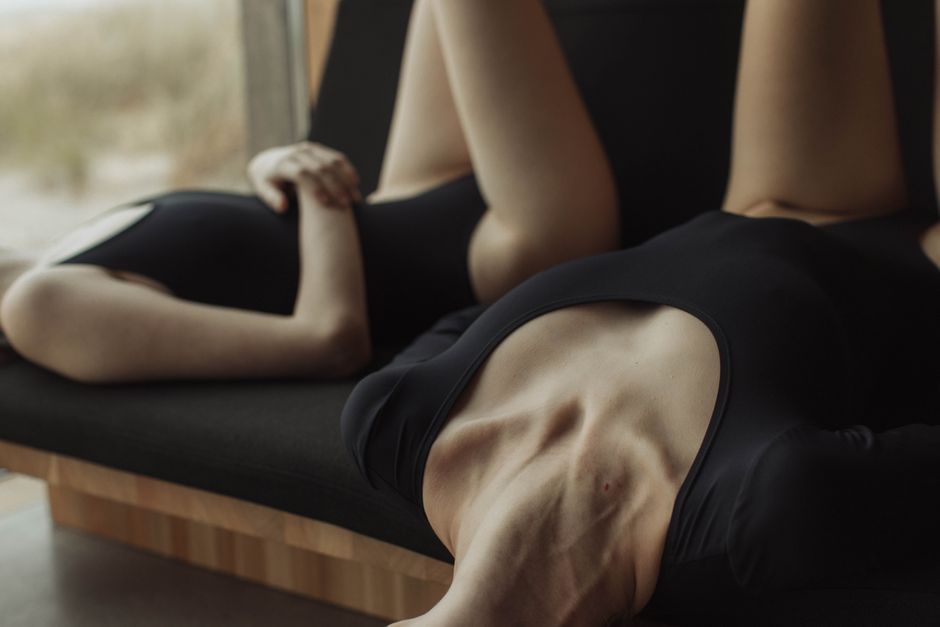 Two women in black swimsuits relax on a sofa indoors, creating a serene and intimate atmosphere.