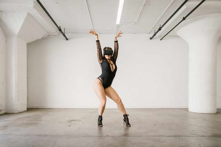 A woman wearing a black leotard and VR headset performs a dance pose indoors.