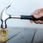 Decentralized Decision-Making: Scaling for Operational Success A hand holding a hammer above a Bitcoin coin on a wooden surface, symbolizing cryptocurrency volatility.