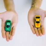 Pair of adult hands holding green and yellow toy cars on a white surface, symbolizing choice.
