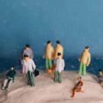 Miniature people figurines on textured surface with blue backdrop, maintaining a whimsical still life theme.