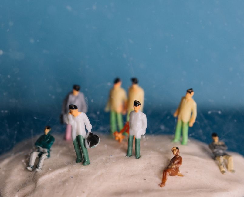 Miniature people figurines on textured surface with blue backdrop, maintaining a whimsical still life theme.