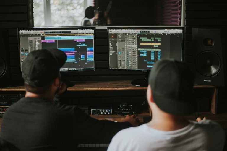 Two men working in a music studio with dual monitors displaying audio editing software.