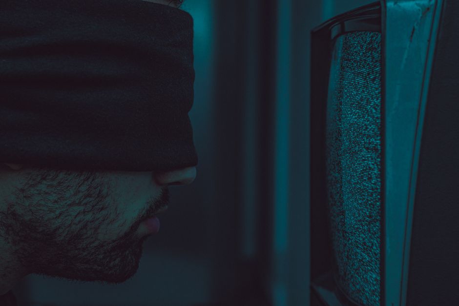 A close-up of a blindfolded man facing a static television screen in a dark room.